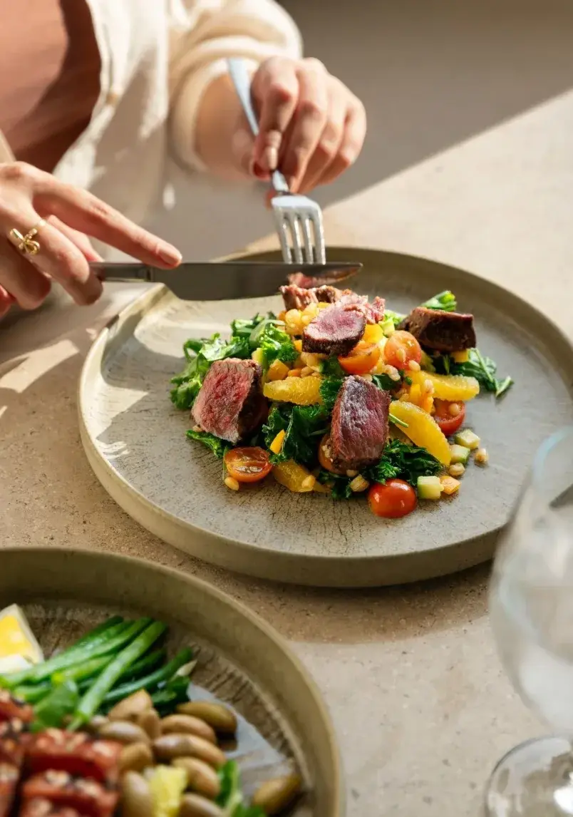 Una persona disfruta de un plato con carne, verduras y cítricos en una mesa de madera.
